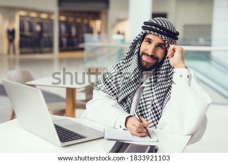 A man sits in a kandura and looks at the camera while writing. A man in a kandura looks at the camera and works at a table.