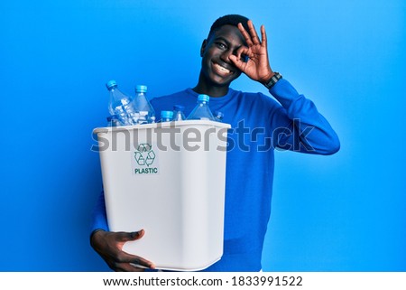 Young african american man holding recycling wastebasket with plastic bottles smiling happy doing ok sign with hand on eye looking through fingers 