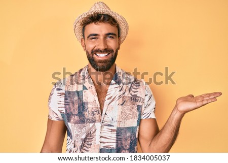 Young hispanic man wearing summer hat smiling cheerful presenting and pointing with palm of hand looking at the camera. 