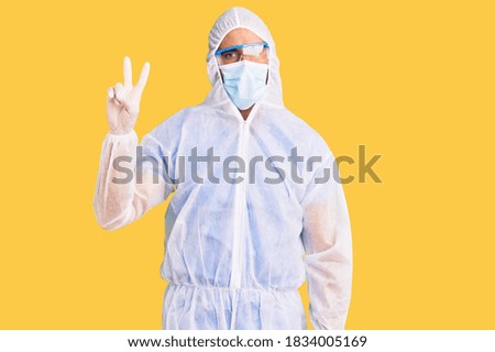 Young hispanic man wearing doctor protection coronavirus uniform and medical mask showing and pointing up with fingers number two while smiling confident and happy. 