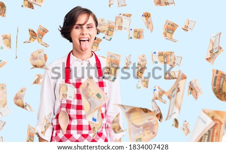 Beautiful young woman with short hair wearing professional cook apron sticking tongue out happy with funny expression. emotion concept.