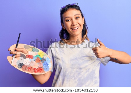 Young woman holding paintbrush and palette smiling happy and positive, thumb up doing excellent and approval sign 