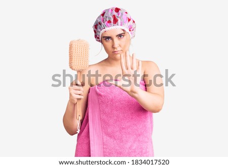 Young caucasian woman wearing shower cap and towel holding sponge with open hand doing stop sign with serious and confident expression, defense gesture 