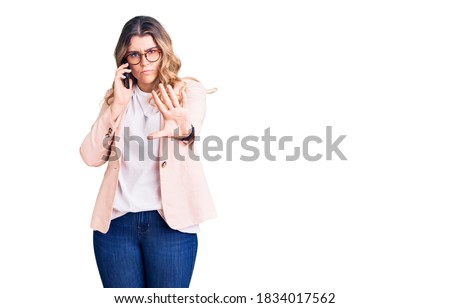 Young caucasian woman having conversation talking on the smartphone with open hand doing stop sign with serious and confident expression, defense gesture 