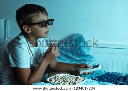 a boy in 3D glasses watches a movie at home at night with a console in his hand and eats popcorn