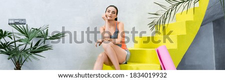 Panoramic orientation of sportswoman sitting on stairs near fitness mat at home
