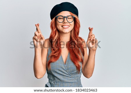 Young redhead woman wearing fashion french look with beret gesturing finger crossed smiling with hope and eyes closed. luck and superstitious concept. 