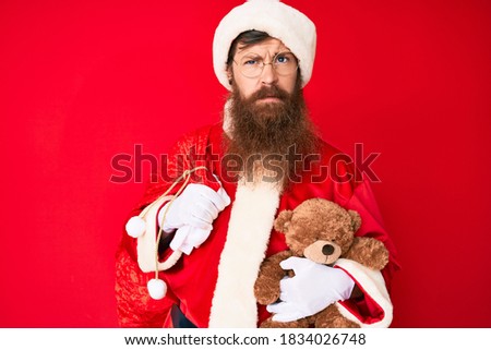 Handsome young red head man with long beard wearing santa claus costume holding teddy bear skeptic and nervous, frowning upset because of problem. negative person. 