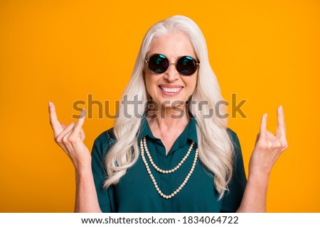 Hard rock Photo of pretty crazy cheerful white haired grandma aged lady showing finger horns metal music lover wear green shirt sun specs necklace isolated bright yellow color background