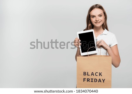 joyful young woman holding digital tablet with blank screen near shopping bag with black friday lettering on grey