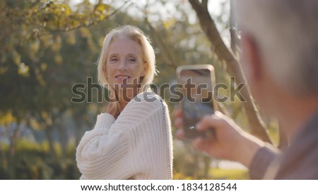 Male pensioner taking picture of attractive senior woman on smartphone in park