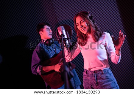 Pretty Asian female singer recording songs by using a studio microphone and pop shield on mic with male playing guitar in blue and red light. Performance and show in the music business. Duet session.