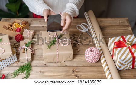 Woman s hands wrapping Christmas gift, close up and taking pictures on the phone. Unprepared presents on wooden background with decor elements and items, top view. New year DIY packing Concept