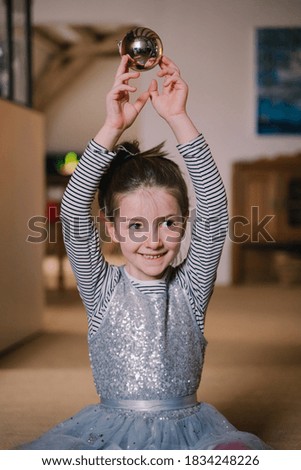 Vertical view of little girl holding a spherical Christmas ornament to hang from a tree at home. Family Christmas concept