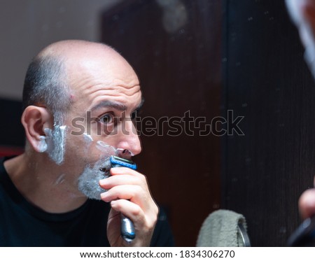 A middle-aged Caucasian man is shaving himself with a hand razor. Fix your gaze on the mirror where you rest the razor