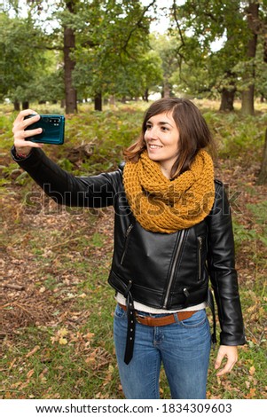 Attractive young female with a black leather jacket and taking a selfie with her smartphone while walking on the park during autumn