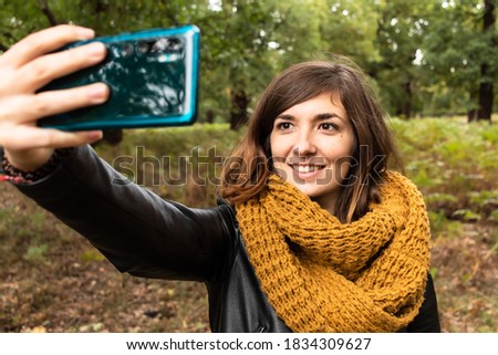 Attractive young female with a black leather jacket and taking a selfie with her smartphone while walking on the park during autumn