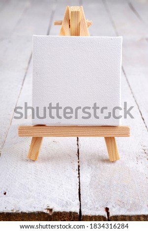 Small easel with white blank cloth on a wooden table. Miniature painting accessories. Light background.