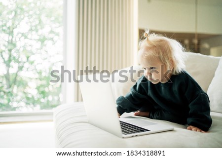 little smiling blonde girl with a ponytail. sitting on the sofa near the monitor, children's clothing. plays educational smart children's games cartoons in the app on the website on your computer lapt