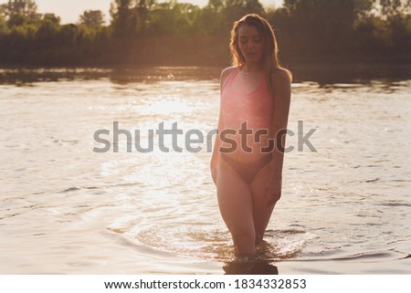 Portrait of young woman swimsuit is standing in the water in a forest lake in the sunset.