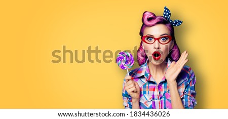 Purple head excited very surprised woman with lollipop. Pinup girl in red glasses, with wide opened mouth, eyes. Beauty model at retro and vintage ad concept. Yellow orange background with copy space 