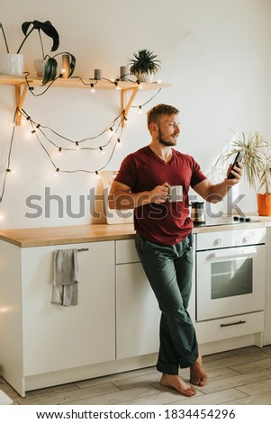 A man of European appearance drinking coffee in a white kitchen. Beautiful kitchen interior. Watching your phone in the morning. High quality photo