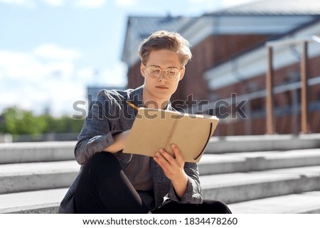 leisure, education and people concept - young man or teenage boy in glasses with notebook, diary or sketchbook writing or drawing in city Royalty-Free Stock Photo #1834478260