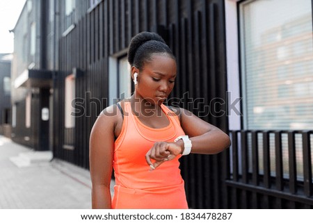 fitness, sport and technology concept - young african american woman with earphones listening to music on smart watch in city