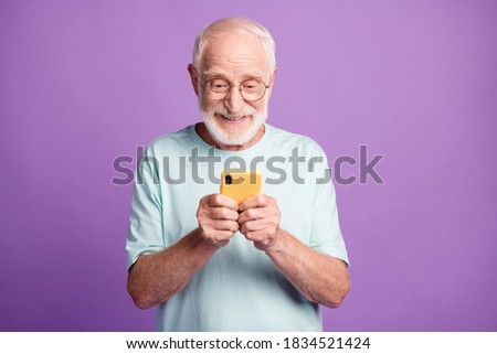 Photo portrait of engaged elderly man holding phone in two hands isolated on vivid purple colored background Royalty-Free Stock Photo #1834521424