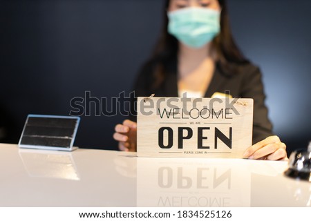 Business owner attractive young Asian woman in suitcase we're open sign on front counter welcoming clients to new hotel.Happy waiter with protective face mask holding open sign while stand at hotel .