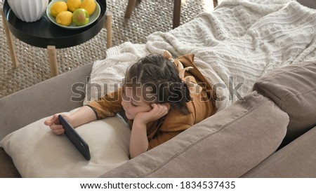 Cute little girl with bows covered with white plaid watches cartoons on cellphone lying on sofa in light living room closeup