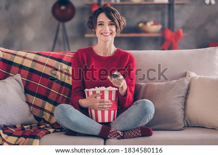 Photo of charming young lady hold remote control popcorn bucket sitting couch wear red pullover jeans socks indoors