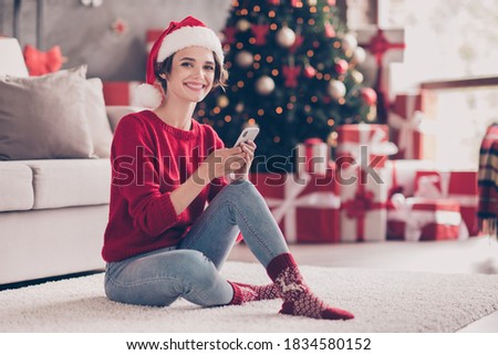 Photo of cute young girl sit carpet hold smartphone look camera wear santa headwear red pullover jeans socks decorated living room indoors