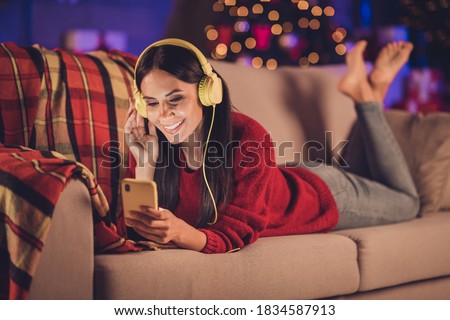 Photo portrait of girl laying on sofa touching earphones holding phone in one hand wearing red jumper indoors with christmas decorations