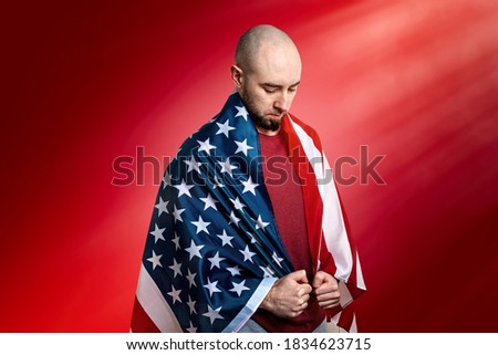 A Caucasian man covered with an American flag stands in a humble and proud pose. Red background. Copy space. Concept of memorial Day, independence day, and other national holidays in the United States