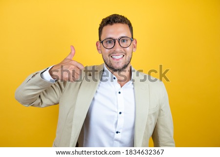 Young handsome businessman wearing suit over isolated yellow background doing happy thumbs up gesture with hand. Approving expression looking at the camera with showing success