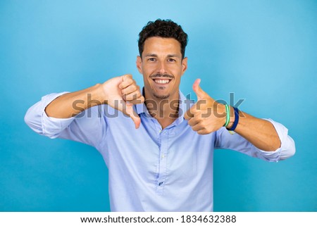 Young handsome man wearing blue shirt over isolated blue background Doing thumbs up and down, disagreement and agreement expression. Crazy conflict