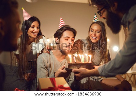 Handsome young man in his twenties, wearing a funny party hat, blowing burning candles on his birthday cake and making a wish, surrounded by happy, smiling friends who came to the party