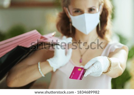 Life during covid-19 pandemic. Closeup on middle aged woman shopper in pink blouse with medical mask, white rubber gloves, credit card and paper shopping bags.