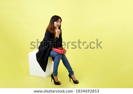 Happy attractive young woman sitting on blank empty box holding red gift box on yellow background