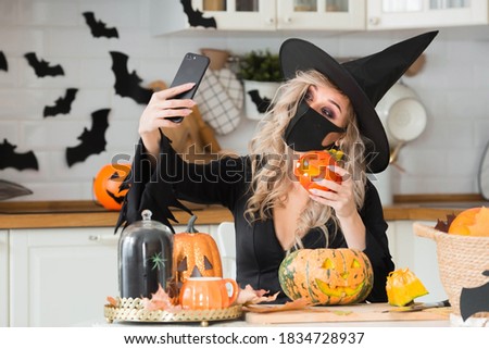Happy Halloween. Online greetings. A woman wearing a black medical mask and a witch costume uses a mobile phone to make video calls to friends and parents.