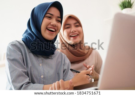 attractive cheerful young muslim business woman working on laptop and smiling while sitting at her desk modern office with her friend at studio