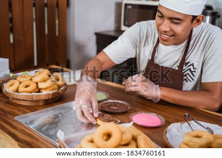 The male chef gives chocolate to decorate donuts in the kitchen, making homemade donuts Royalty-Free Stock Photo #1834760161
