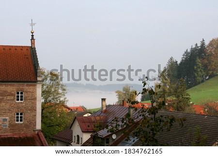 Pastel views of the mountain landscape with roofs of old buildings, fog, meadow and green trees on a cloudy day in the Allgäu region, just.