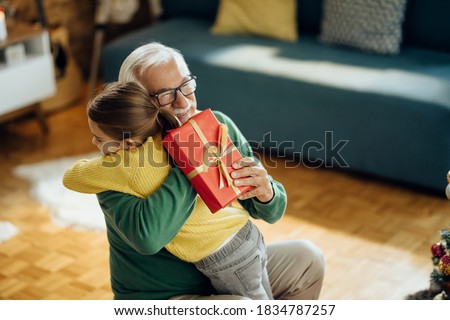 Happy senior man embracing his granddaughter while receiving a gift on Christmas at home.  Royalty-Free Stock Photo #1834787257