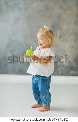 little girl blonde short hair, in a white t-shirt and jeans, stands and holds a glass of water