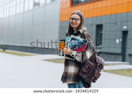 Portrait of girl student standing at university campus. Laughing Woman wearing braces, eyeglasses, bag with books having coffee break after lecture. Enjoying College life. Learning education concept. Royalty-Free Stock Photo #1834814329