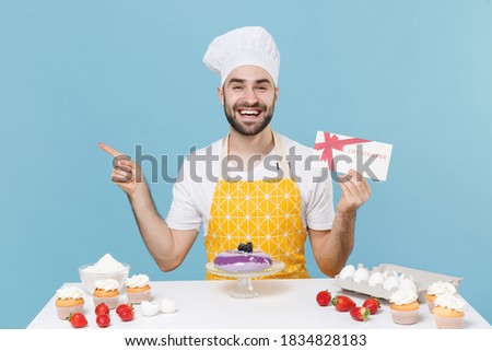 Cheerful young male chef or cook baker man in apron white t-shirt toque chefs hat cooking at table isolated on blue background. Cooking food concept. Hold gift certificate pointing index finger aside