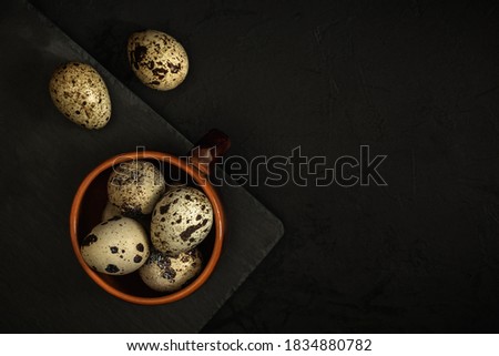 raw spotted quail eggs in a brown clay cup on the edge of a black stone cutting board on a textured dark concrete surface. moody artistic mockup with copy space. top view