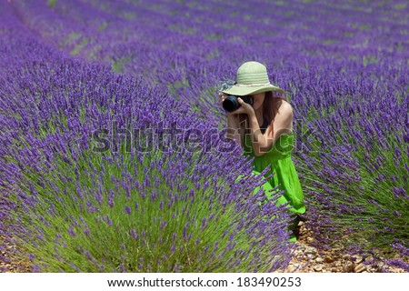 Beautiful woman among lavender photographing some lavender flowers. Wearing green dress and hat, decorated with lavender twigs. Placed in foreground, side view.